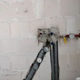 Surowy mur z widocznymi rurami instalacji wodnej, z czerwonymi i niebieskimi zakończeniami, oraz manometrem z żółtym zaworem, w trakcie montażu.