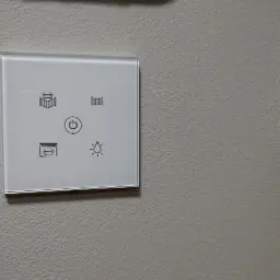 Szklany panel dotykowy na ścianie, z ikonami sterowania bramą wjazdową, garażową, furtką, włącznikiem i regulacją oświetlenia.