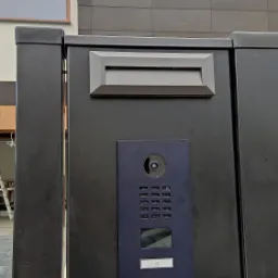 Czarny panel bramy z wbudowanym wideodomofonem DoorBird, skrzynką na listy i numerem domu '9', na tle nowoczesnego domu z szarą elewacją.