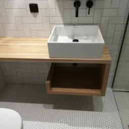 Nowoczesna łazienka: biała, kwadratowa umywalka nablatowa na blacie z jasnego drewna, czarna armatura, białe kafelki ułożone w cegiełkę oraz mozaika na podłodze.