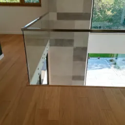 Jasna, drewniana podłoga z widokiem na szklaną balustradę schodów i okno z widokiem na ogród, ściana wykończona szarymi płytkami w kwadraty.