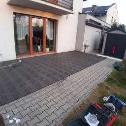 Artis Project - Montaż modułowego tarasu z kompozytu w kolorze grafitowym, widoczne narzędzia i materiały budowlane, częściowo ułożona nawierzchnia przy domu.