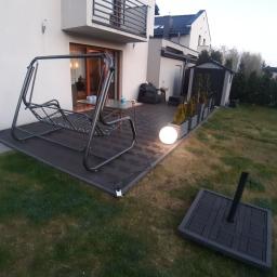 Artis Project - Nowoczesny taras z ciemnej kostki brukowej z huśtawką ogrodową i kulistą lampą, widok na budynek mieszkalny i zadbany ogród.