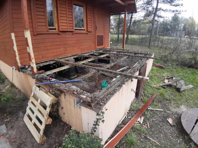 Demontaż starego drewnianego tarasu przy brązowym domku, widoczne narzędzia, paleta i elementy konstrukcji tarasu w trakcie rozbiórki.