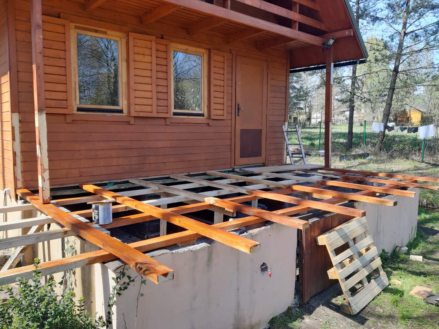 Konstrukcja drewnianego tarasu w trakcie budowy przy domku letniskowym, widoczne legary pomalowane impregnatem, puszka z impregnatem na legarach, drabina oparta o ścianę domku.