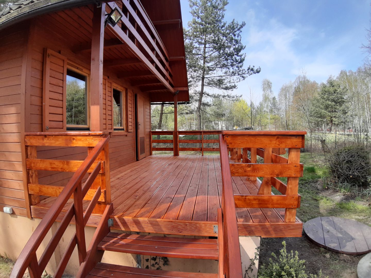 Drewniany taras przy domku letniskowym z balustradą i schodami, widok na las w tle, jasne niebo.