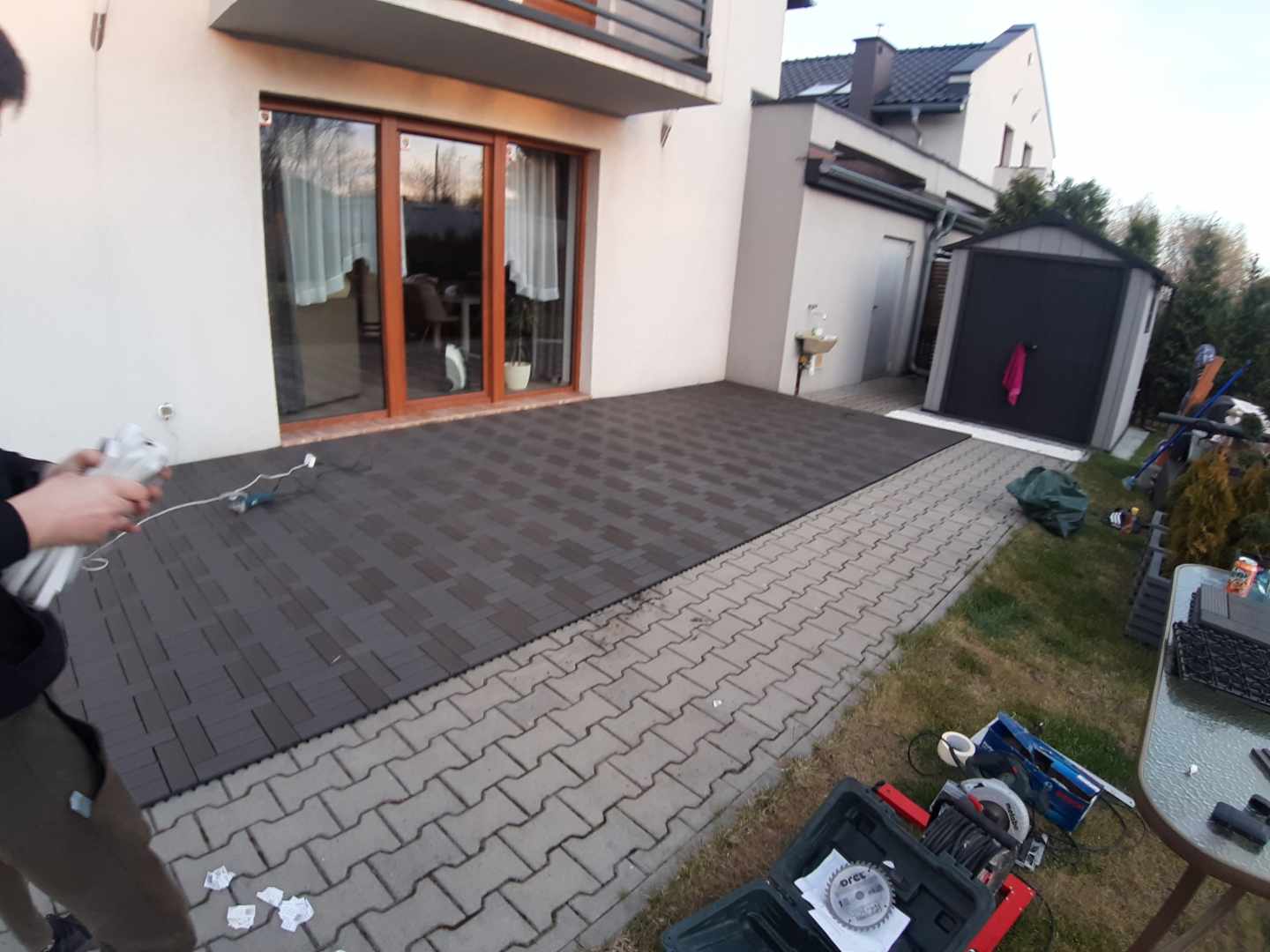 Montaż modułowego tarasu z kompozytu w kolorze grafitowym, widoczne narzędzia i materiały budowlane, częściowo ułożona nawierzchnia przy domu.