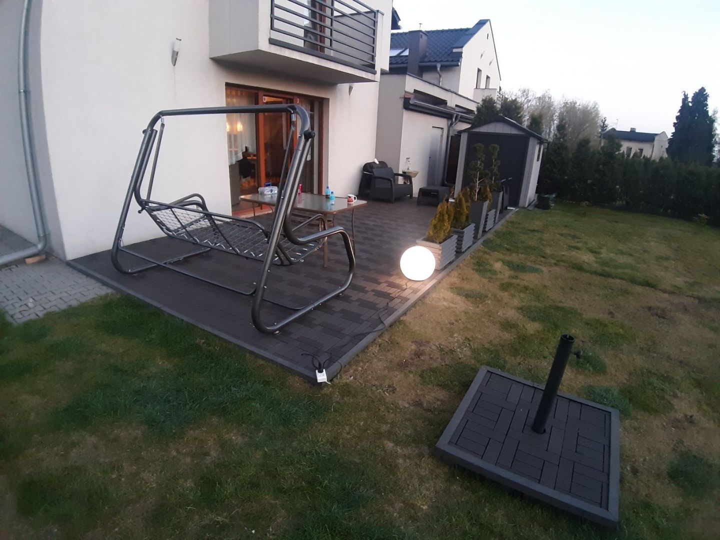 Nowoczesny taras z ciemnej kostki brukowej z huśtawką ogrodową i kulistą lampą, widok na budynek mieszkalny i zadbany ogród.