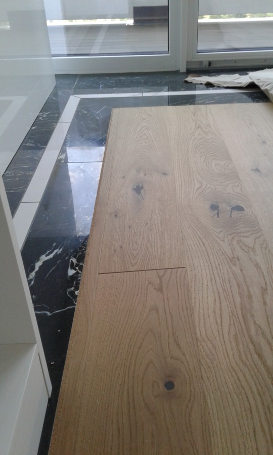 Fragment dębowej deski warstwowej z widocznymi słojami i sękami, ułożonej obok błyszczącej, czarnej płytki marmuropodobnej z białymi żyłami i białą obwódką, widok z góry.