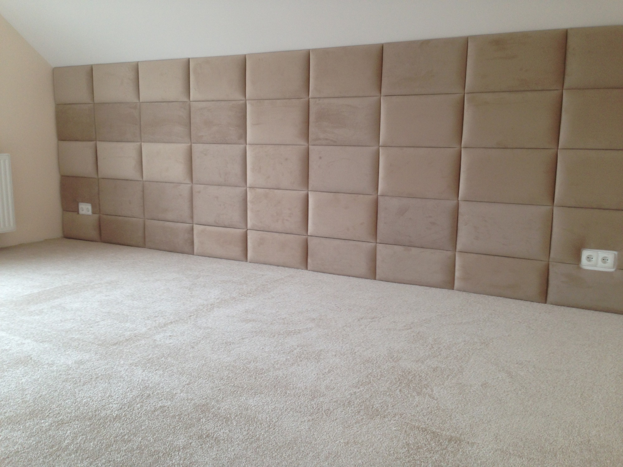 Sypialnia z beżową wykładziną dywanową i tapicerowanym panelem ściennym w kwadraty, widoczne gniazdka elektryczne.