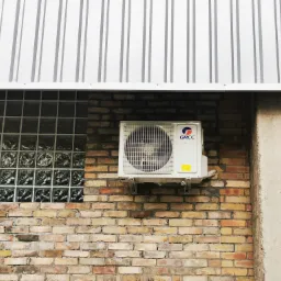 Biała jednostka zewnętrzna klimatyzatora marki Gree zamontowana na ceglanej ścianie budynku z oknem z luksferów i metalowym zadaszeniem.