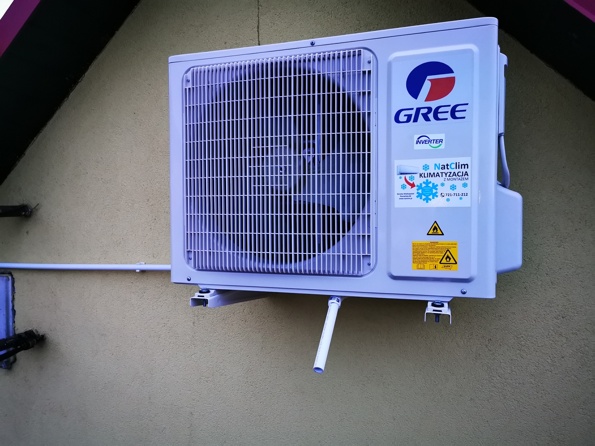 Zewnętrzna jednostka klimatyzacji marki Gree, zamontowana na elewacji budynku z widocznym odprowadzeniem skroplin i naklejką informacyjną.