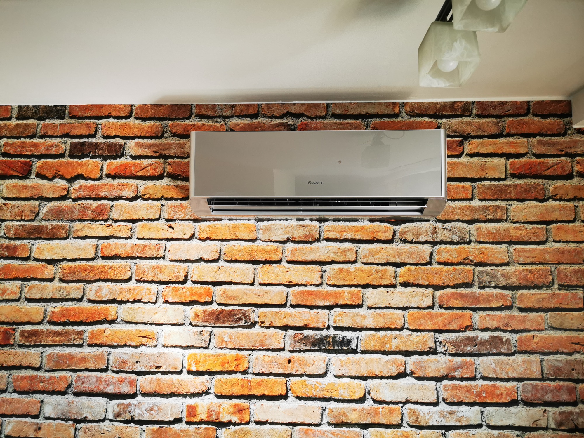 Szary klimatyzator marki Gree zamontowany na ścianie z czerwonej cegły w pomieszczeniu z białym sufitem i lampą