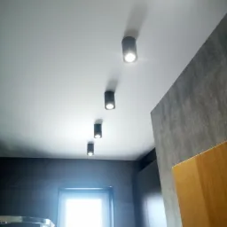 Rząd czterech czarnych, cylindrycznych lamp sufitowych LED w nowocześnie wykończonym wnętrzu z szarą ścianą i oknem. Widok z dołu.