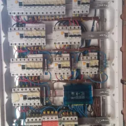 Rozdzielnica elektryczna z zabezpieczeniami różnicowoprądowymi i nadprądowymi, okablowanie w kolorach niebieskim, czerwonym i brązowym, zamontowana na ścianie z cegły.