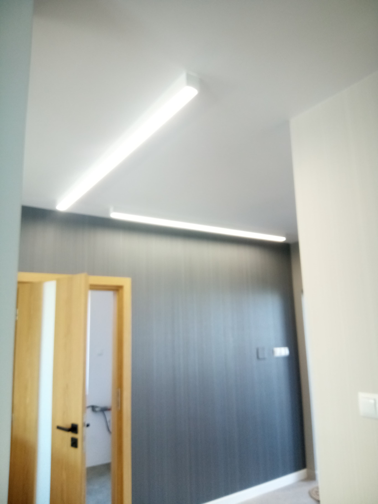 Dwie liniowe lampy LED zamontowane na suficie wzdłuż ściany z szarą tapetą i drewnianymi drzwiami z czarną klamką.