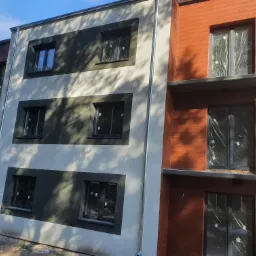 Nowoczesny budynek wielorodzinny z fragmentem elewacji w kolorze terakoty i białym tynkiem, widoczne okna zabezpieczone folią malarską, rynna spustowa.