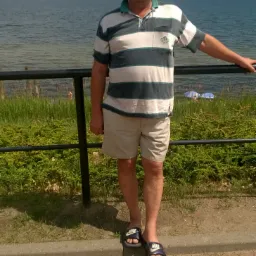 Mężczyzna w okularach przeciwsłonecznych i klapkach Nike stoi na tle morza i nieba, opierając się o barierkę.