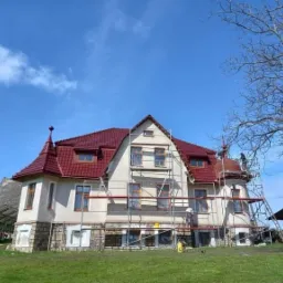 Dom z nowym, czerwonym dachem z dachówki, częściowo otoczony rusztowaniem, w trakcie prac wykończeniowych, widoczne panele słoneczne w tle.