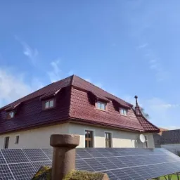 Czerwony dach z dachówki karpiówki na starym domu z oknami w lukarnach, panele słoneczne na pierwszym planie, błękitne niebo w tle.