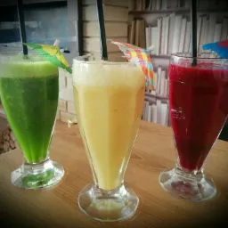 Trzy szklanki z kolorowymi koktajlami: zielonym, żółtym i czerwonym, ozdobione małymi parasolkami i słomkami, ustawione na drewnianym stole na tle regału z książkami.