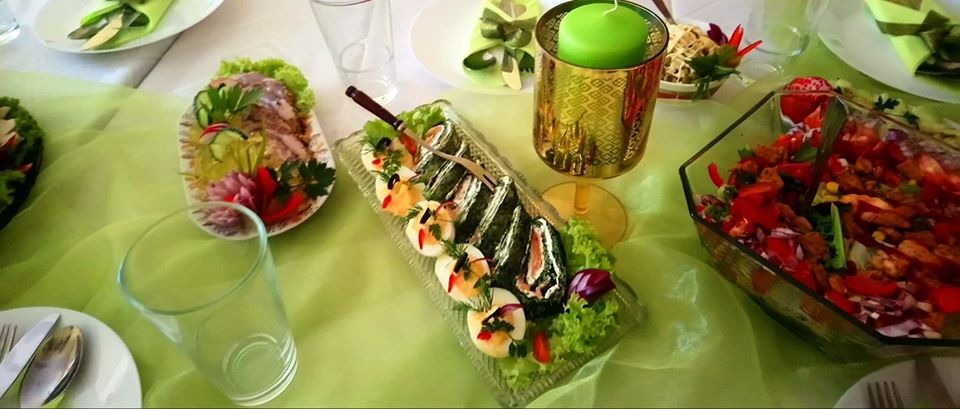 Dekoracyjnie zastawiony stół z różnorodnymi potrawami: sałatkami, roladkami z łososiem i jajkami, śledziem w galarecie, zieloną świecą w ozdobnym świeczniku, całość utrzymana w zielonej tonacji.
