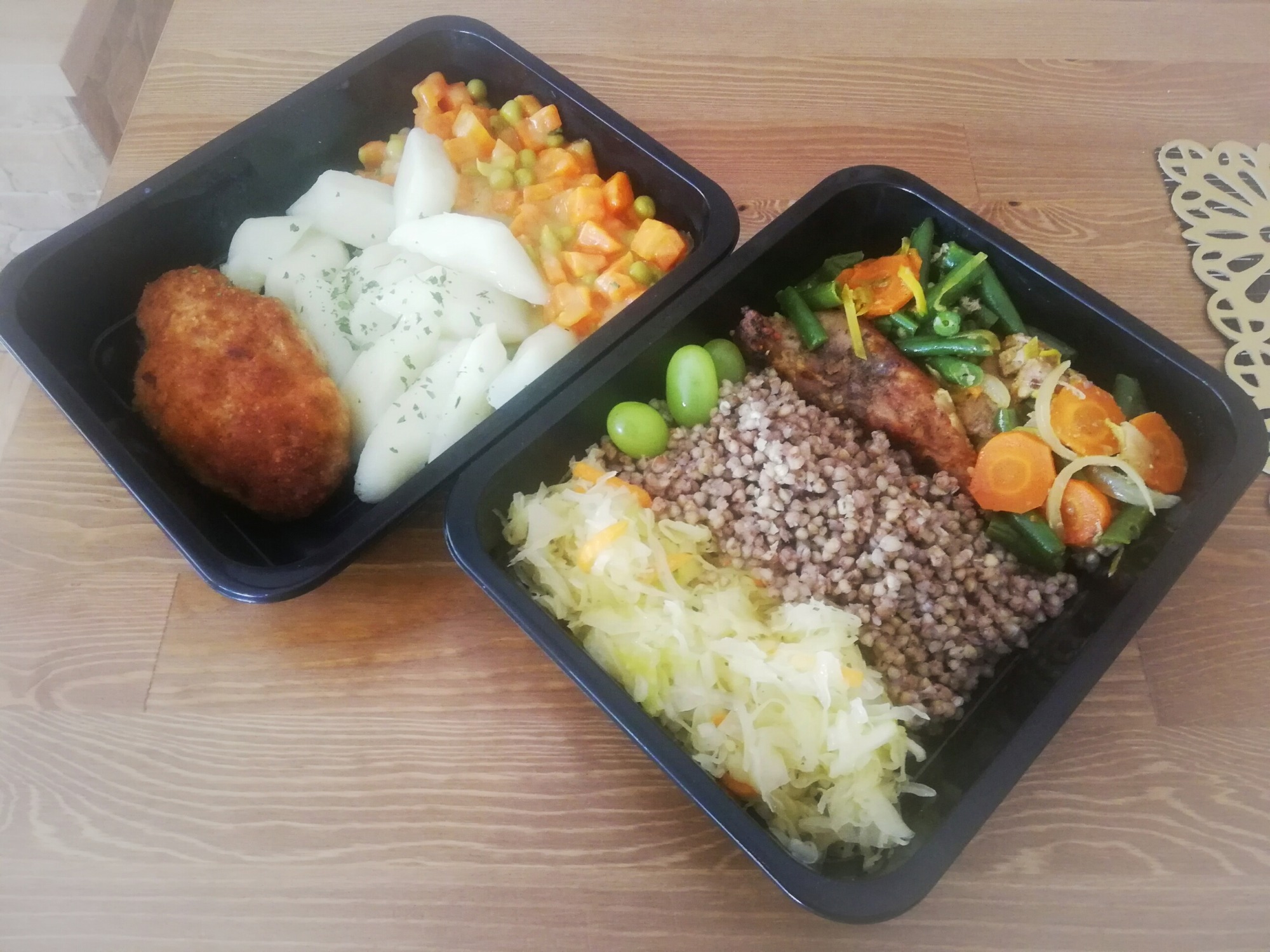 Dwa czarne pojemniki z gotowymi daniami: jeden z kotletem, ziemniakami i warzywami w sosie, drugi z kaszą gryczaną, kapustą kiszoną i warzywami, widoczne trzy zielone winogrona.