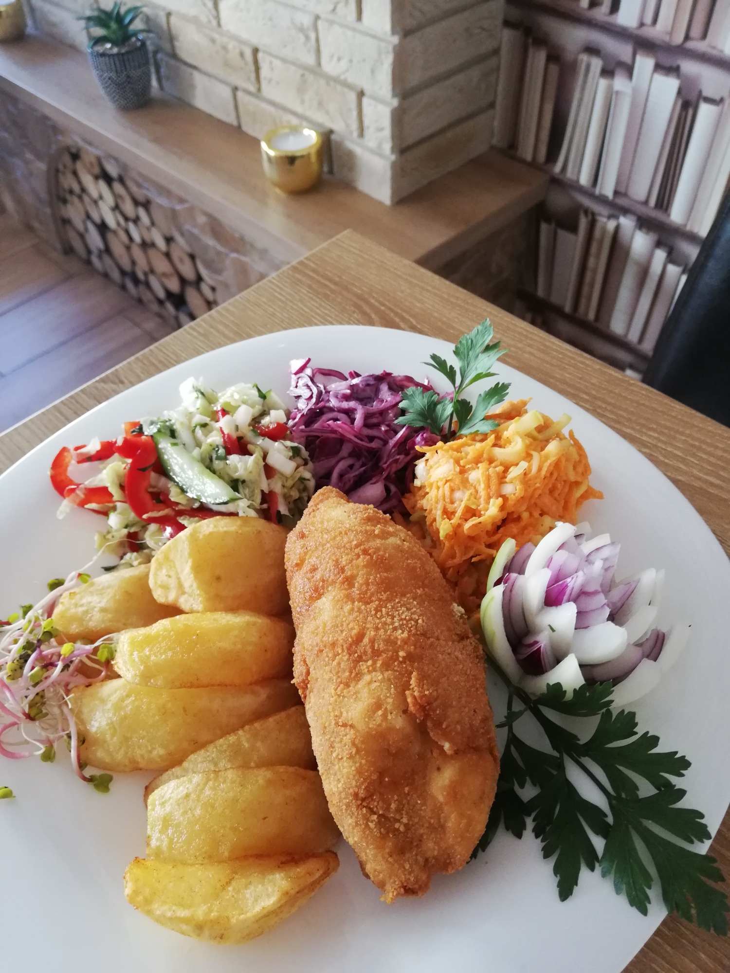Kotlet devolay na białym talerzu, otoczony frytkami, surówkami z kapusty białej i czerwonej, marchewki i dekoracją z czerwonej cebuli oraz natki pietruszki.