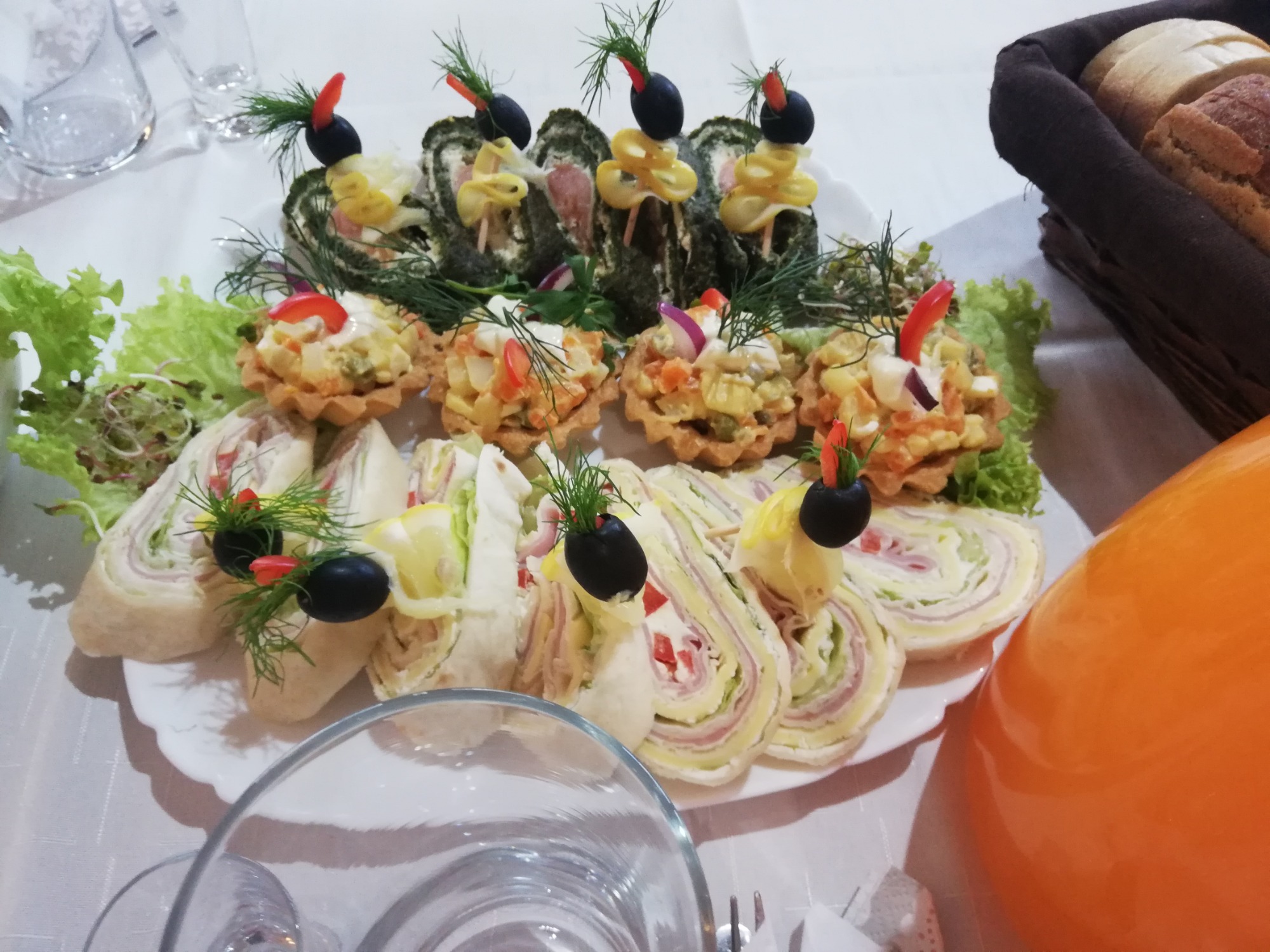 Dekoracyjnie ułożone na białym talerzu przekąski cateringowe: roladki z tortilli z szynką i warzywami, tartaletki z sałatką jarzynową i szpinakowe roladki, ozdobione oliwkami i papryką, widoczne...