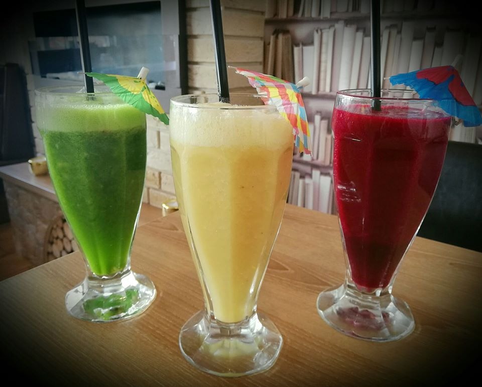 Trzy szklanki z kolorowymi koktajlami: zielonym, żółtym i czerwonym, ozdobione małymi parasolkami i słomkami, ustawione na drewnianym stole na tle regału z książkami.