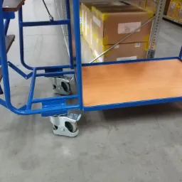 Niebieski wózek magazynowy z drewnianą platformą i schodkami, wyposażony w obrotowe kółka z hamulcami, stojący na betonowej posadzce magazynu.