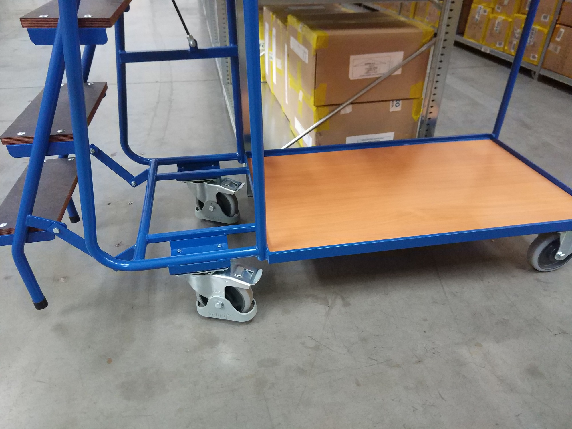 Niebieski wózek magazynowy z drewnianą platformą i schodkami, wyposażony w obrotowe kółka z hamulcami, stojący na betonowej posadzce magazynu.