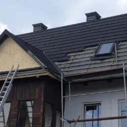Częściowo pokryty dach z oknem dachowym, widoczne łaty, kominy w kolorze grafitowym, rusztowanie i drabina oparta o drewnianą część budynku.