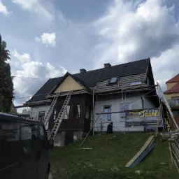 Dom w trakcie wymiany pokrycia dachowego, widoczne rusztowania, drabiny i baner reklamowy firmy dekarskiej, częściowo zdjęta stara dachówka, błękitne niebo z chmurami w tle.