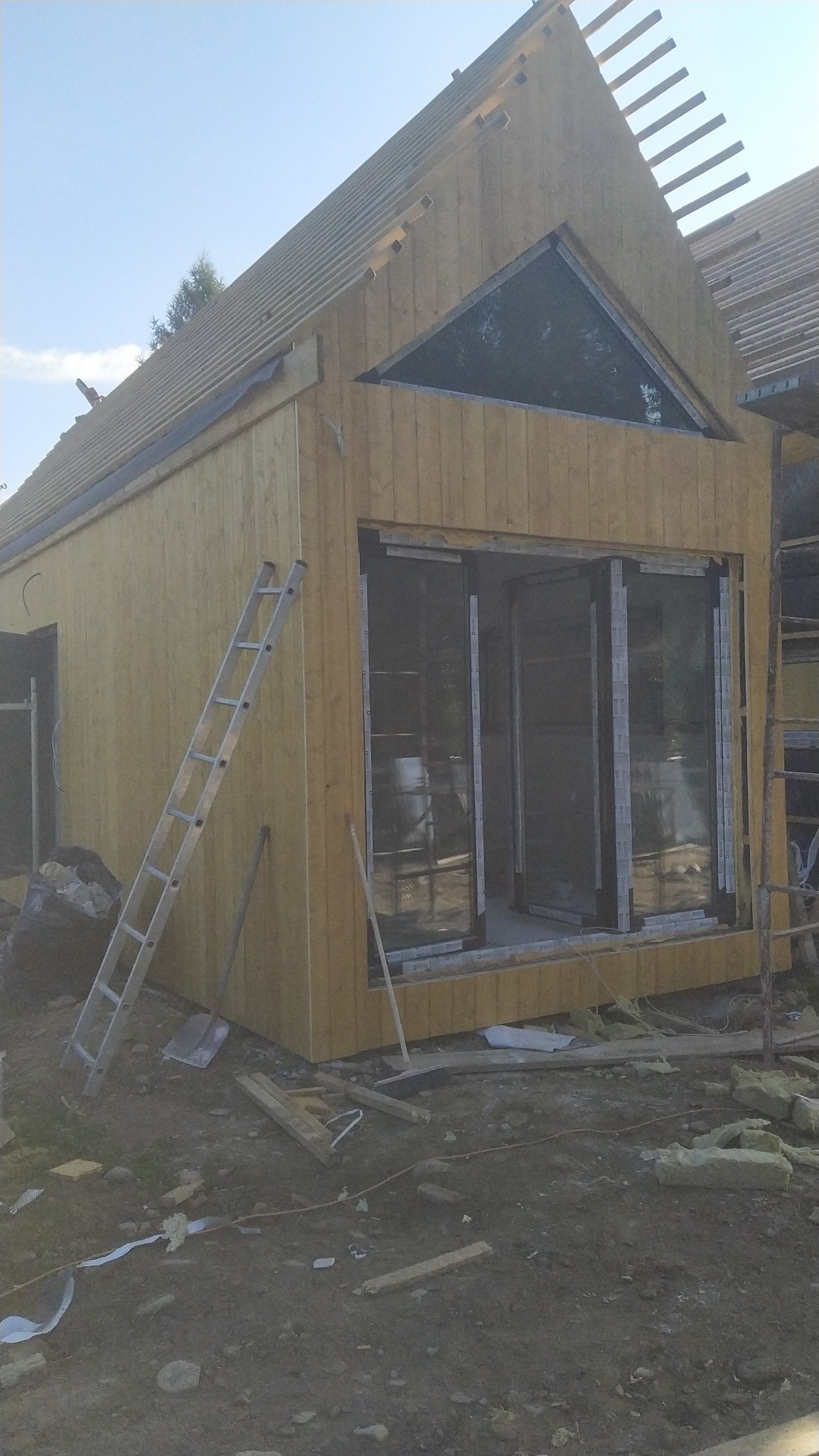 Surowy stan budowy domu z drewnianą elewacją, widoczne rusztowanie dachu, okno trójkątne w szczycie, duże przesuwne okna na parterze, drabina oparta o ścianę, teren budowy z narzędziami i resztkami...