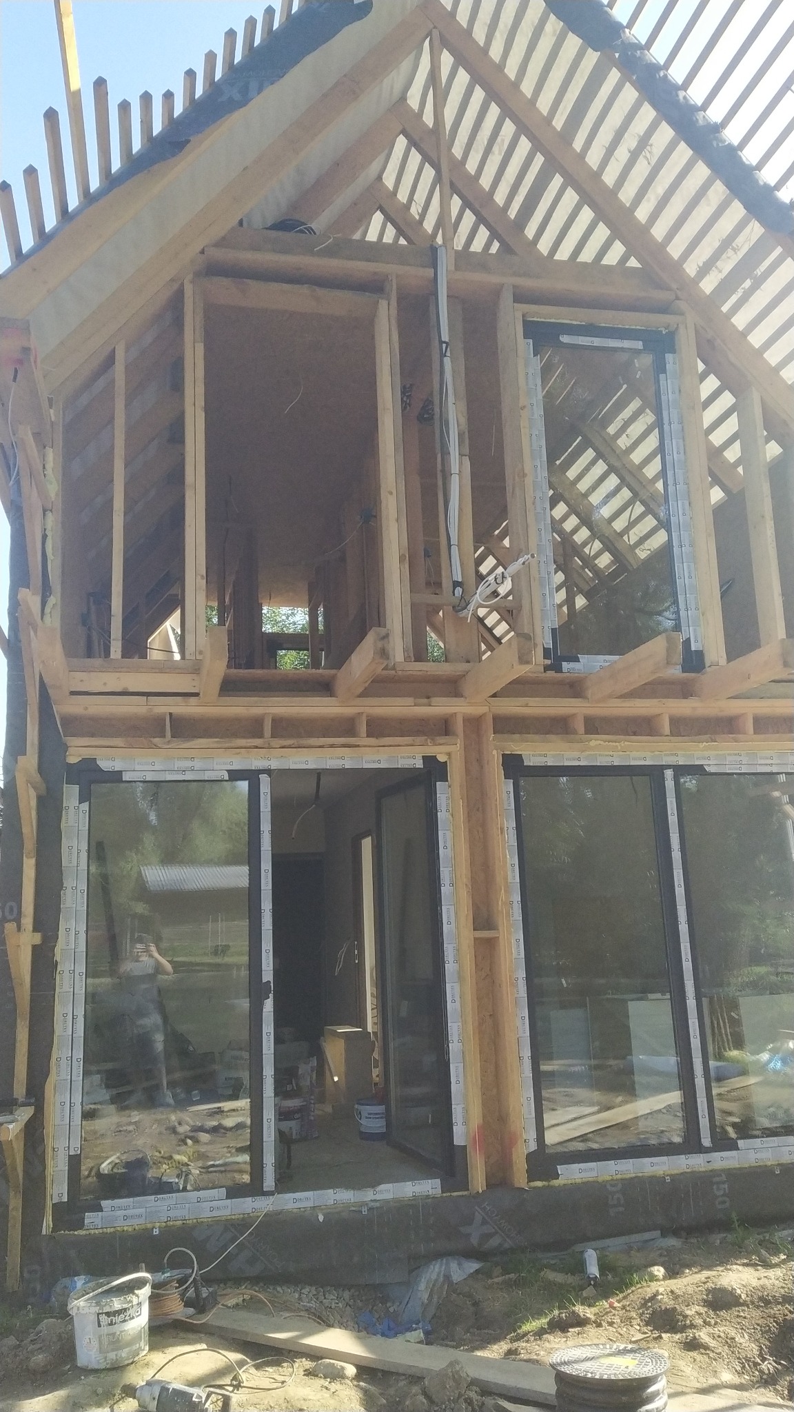 Konstrukcja drewniana domu w trakcie budowy, widoczne okna z czarnymi ramami, elementy izolacji i narzędzia budowlane na ziemi, osoba odbija się w szybie okna.