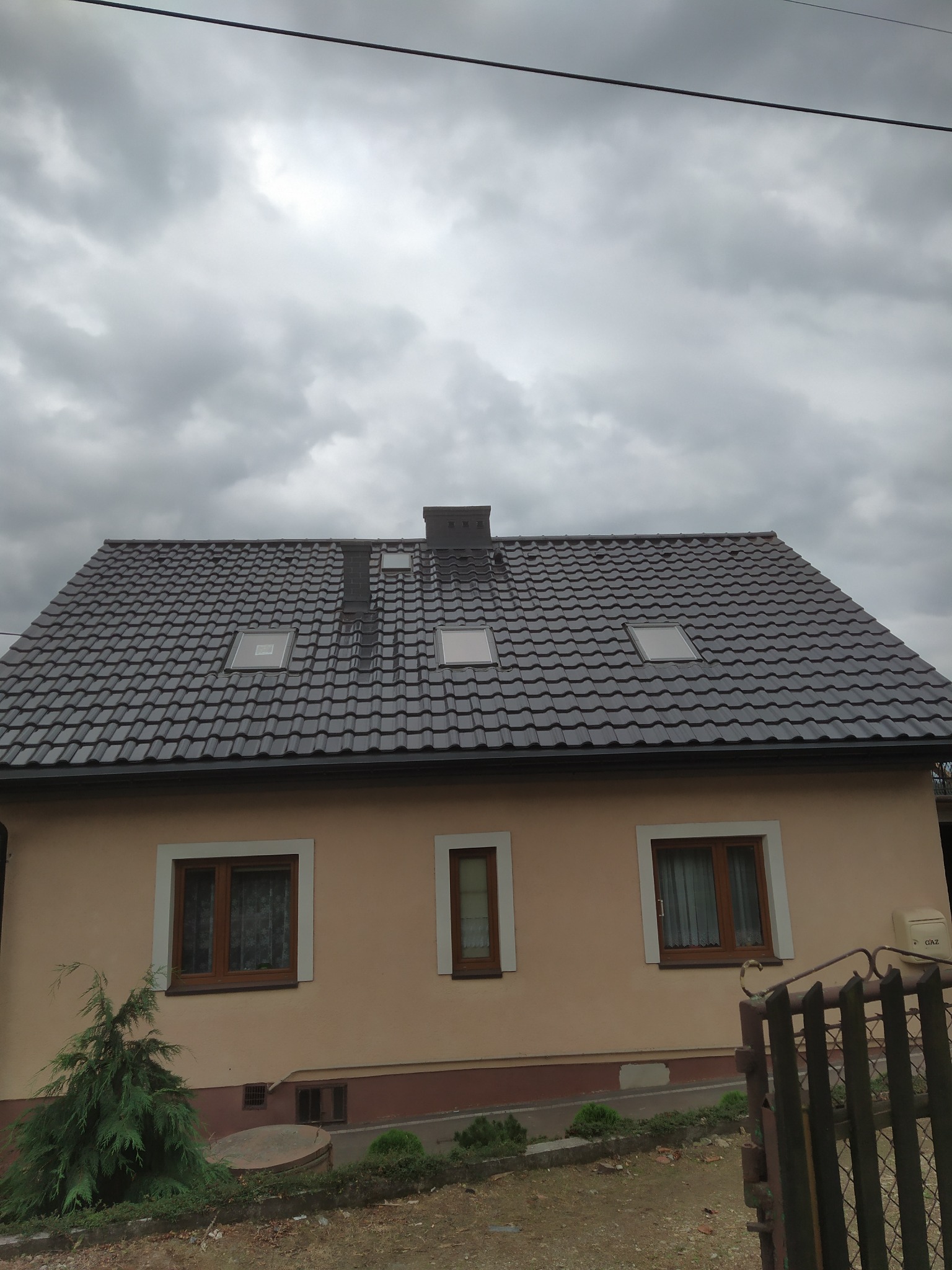 Dom jednorodzinny z nowym, ciemnoszarym dachem z dachówki ceramicznej, widoczne okna dachowe i komin, pochmurne niebo w tle.