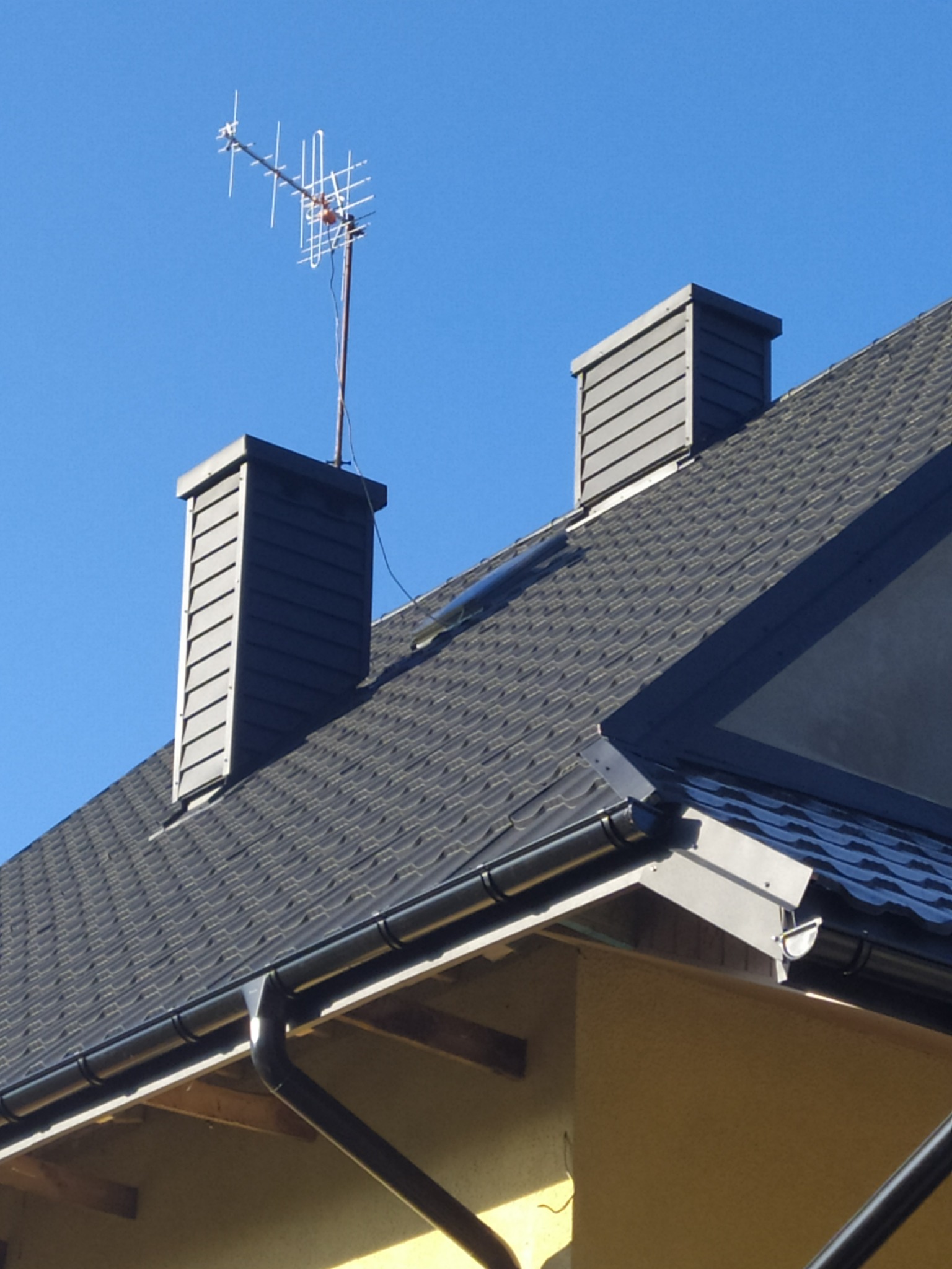 Czarny dach z dachówki bitumicznej, dwa kominy z szarej blachy trapezowej i antena telewizyjna na tle błękitnego nieba.