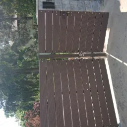 Drewniana brama dwuskrzydłowa z kompozytu, widok z zewnątrz, z widocznymi elementami mocującymi i zamkiem, na tle zieleni i betonowej nawierzchni.