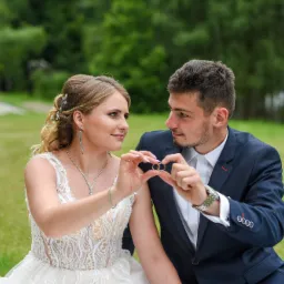 Para narzeczonych trzymająca obrączki ślubne na tle zieleni, panna młoda w koronkowej sukni, pan młody w granatowym garniturze, romantyczne spojrzenie.