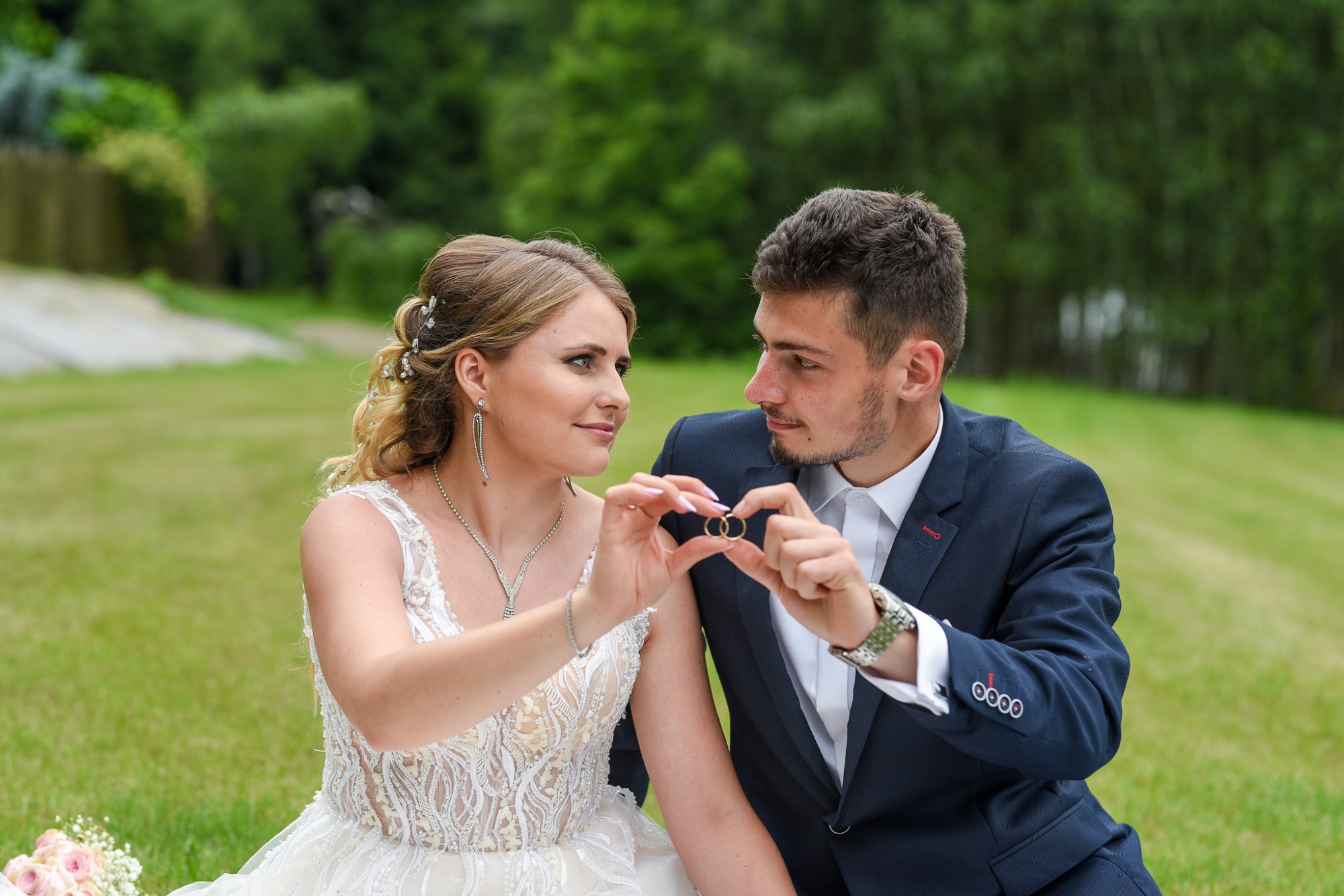 Para narzeczonych trzymająca obrączki ślubne na tle zieleni, panna młoda w koronkowej sukni, pan młody w granatowym garniturze, romantyczne spojrzenie.