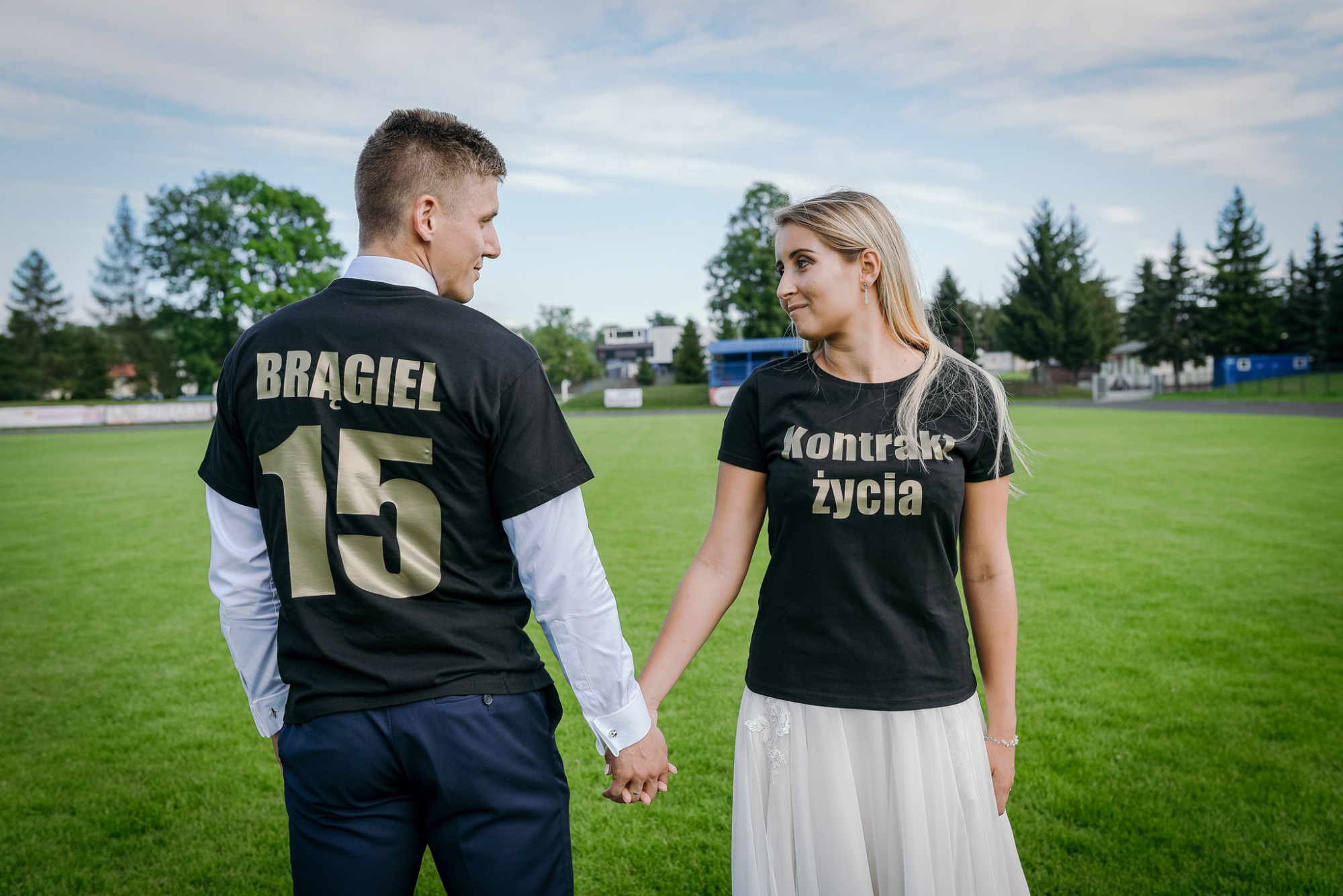 Para trzymająca się za ręce na zielonym boisku sportowym, oboje ubrani w czarne koszulki z nadrukami: mężczyzna z nazwiskiem i numerem, kobieta z napisem 'Kontrakt życia'.