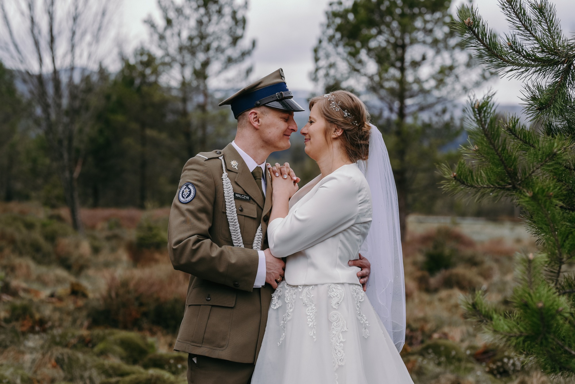 Para młoda: kobieta w białej sukni ślubnej i welonie, mężczyzna w mundurze wojskowym, trzymają się za ręce, patrząc na siebie z czułością na tle leśnego krajobrazu.