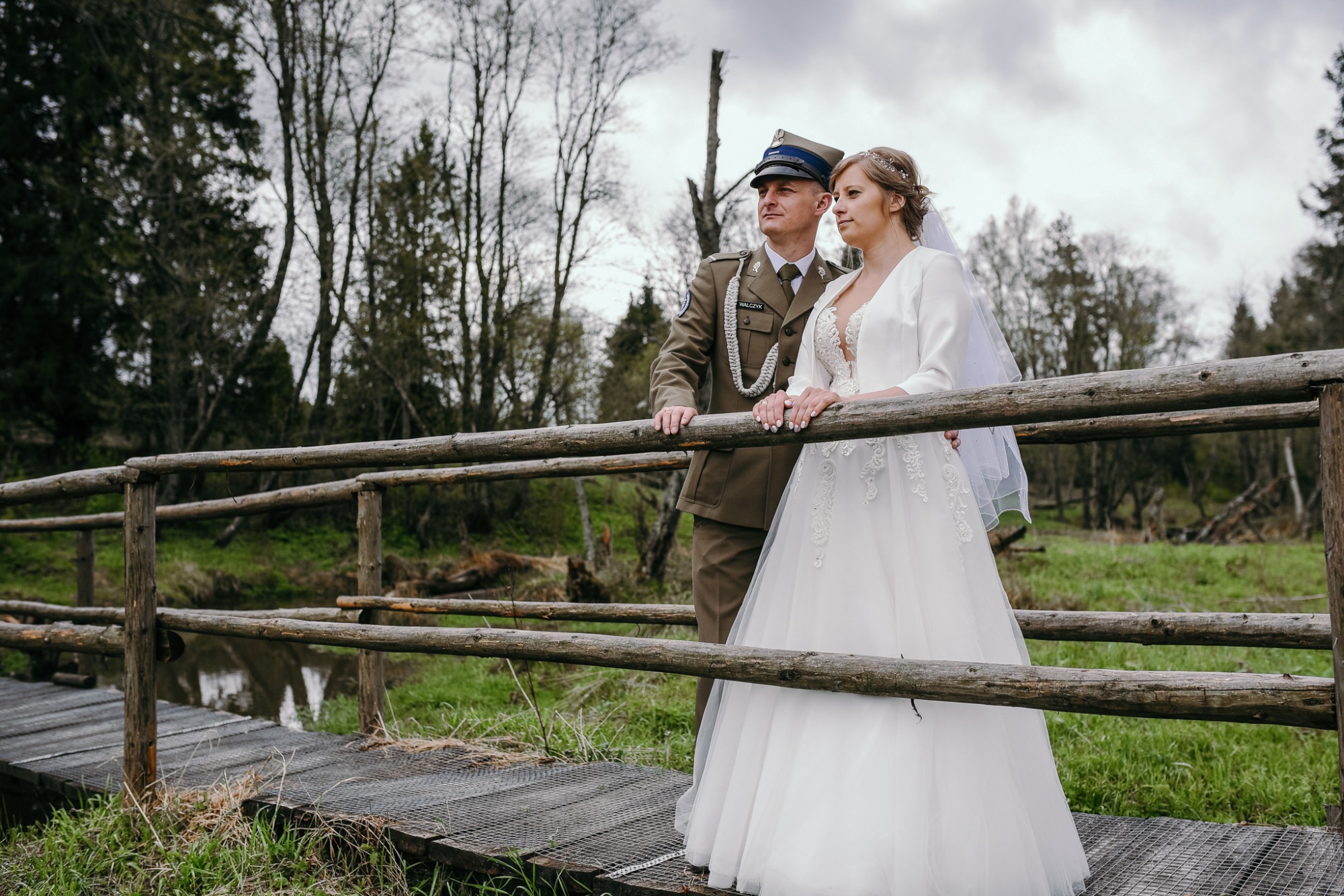 Para młoda na drewnianym mostku w plenerze: pan młody w mundurze Wojska Polskiego, panna młoda w białej sukni ślubnej z welonem, oboje oparci o barierkę mostu, patrzą w dal.