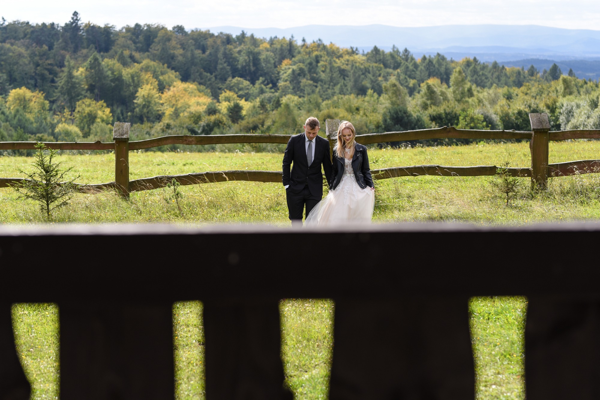 Para młoda spaceruje po łące w sukni ślubnej i czarnej skórzanej kurtce na tle drewnianego płotu i lasu.