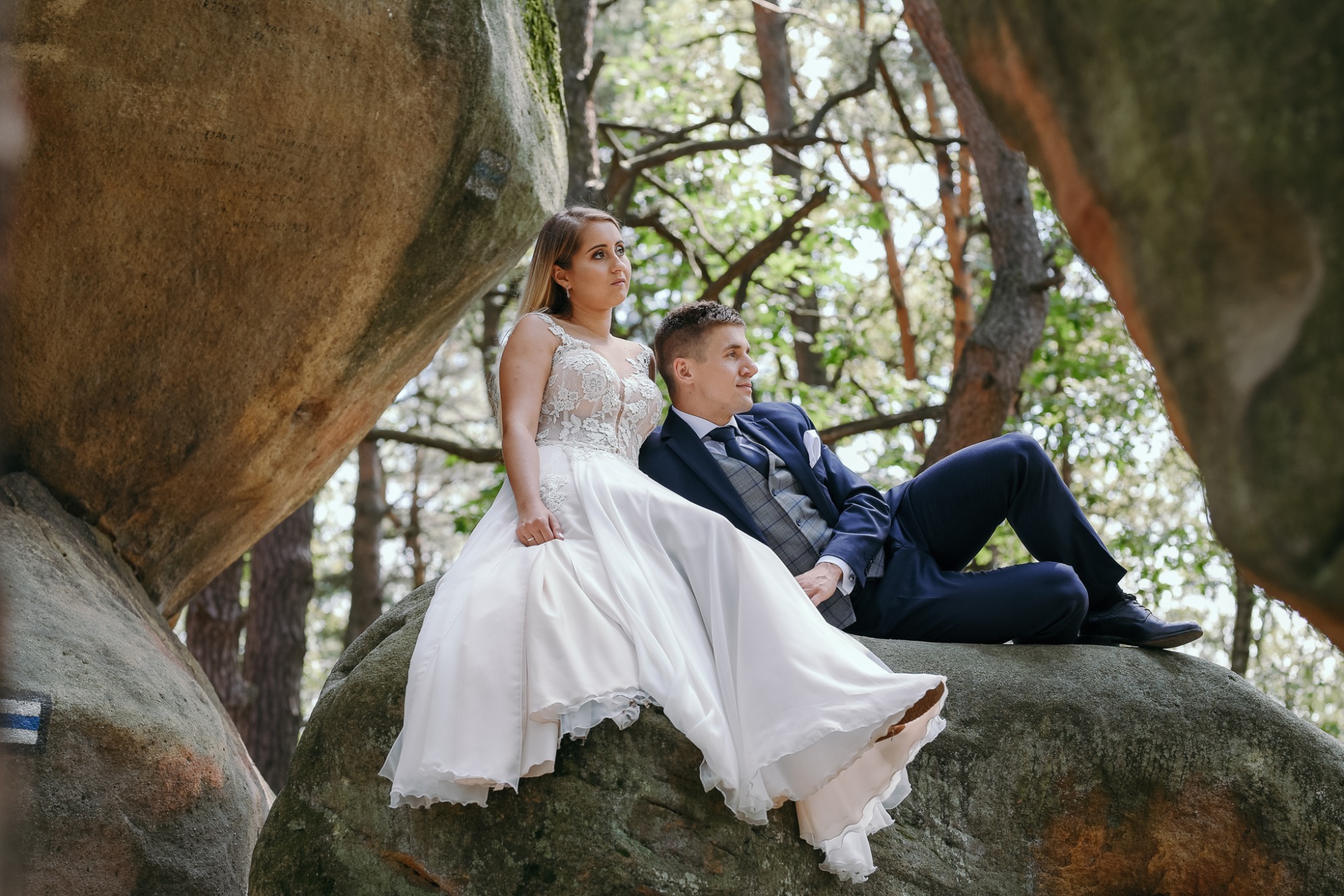 Para narzeczonych w strojach ślubnych pozuje na formacji skalnej w lesie, widoczny szlak turystyczny.