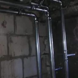 Metalowe rury instalacji hydraulicznej, prowadzone wzdłuż surowej ściany z cegieł, z widocznymi zaworami i mocowaniami.