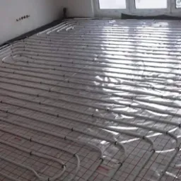 Białe rury ogrzewania podłogowego ułożone na macie grzewczej z siatką w odcieniach czerwieni, widoczne okno dachowe w jasnym pomieszczeniu.