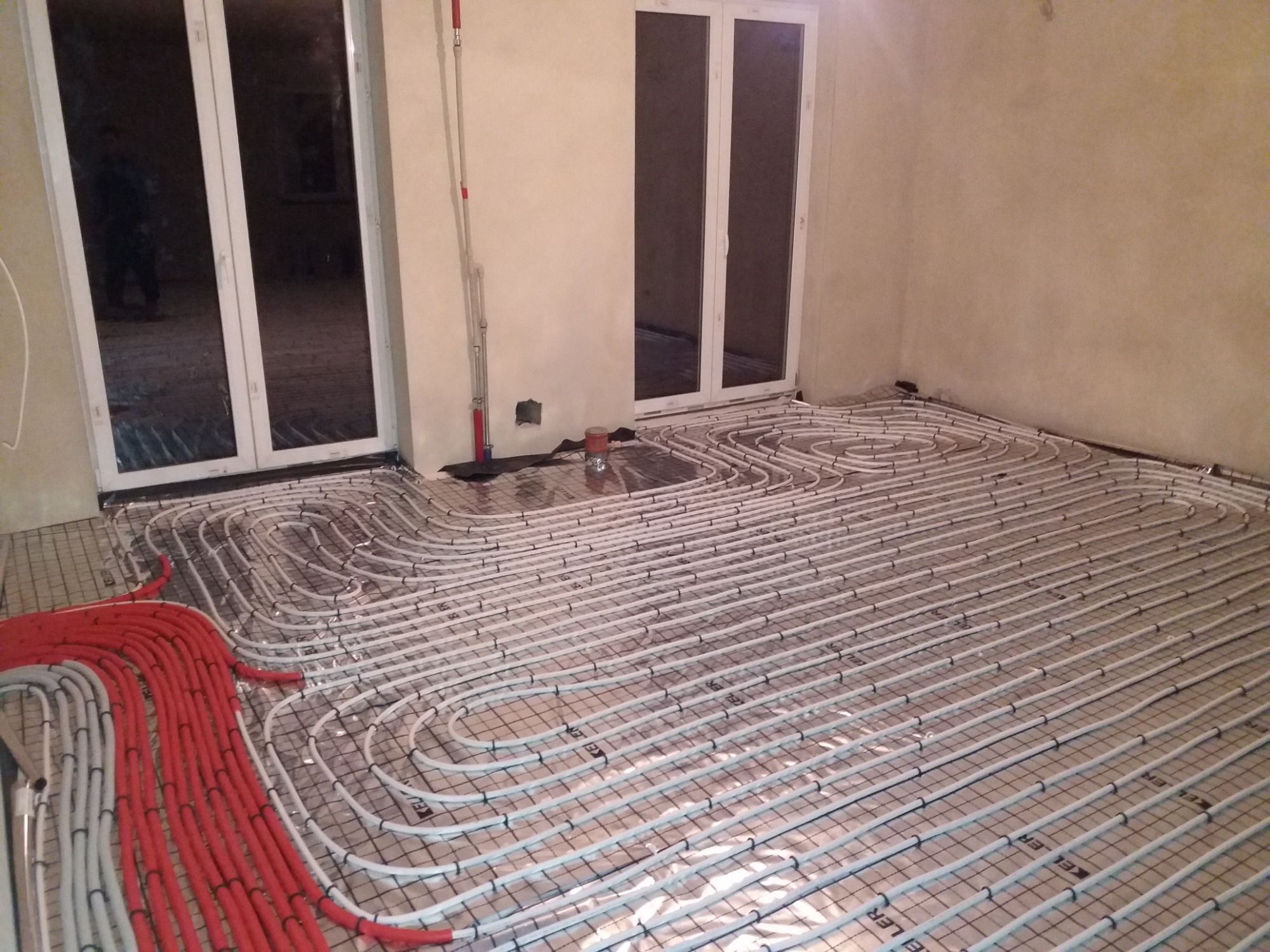 Rozłożone rury ogrzewania podłogowego na folii izolacyjnej z siatką, widoczne czerwone i białe rury, pomieszczenie w stanie surowym z oknami balkonowymi.