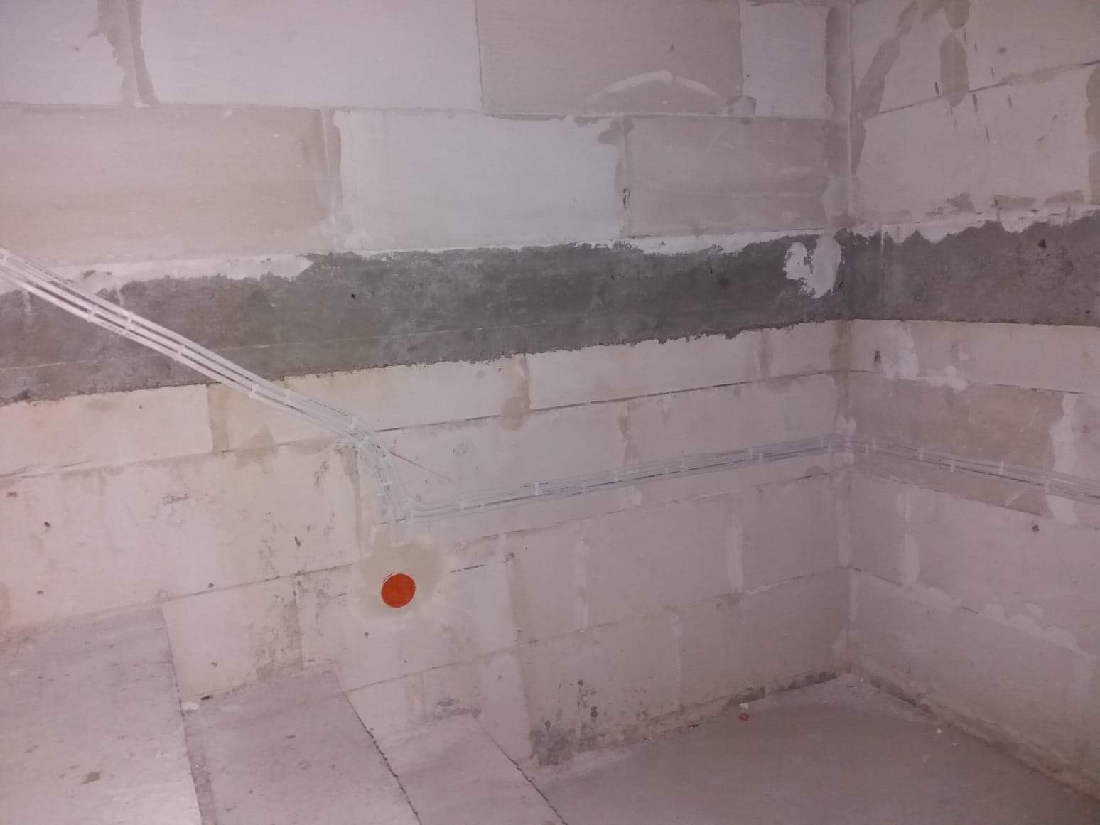 Surowy stan ścian z betonu komórkowego w trakcie prac instalacyjnych, widoczne kable elektryczne prowadzone w peszlach i pomarańczowa puszka pod gniazdko.